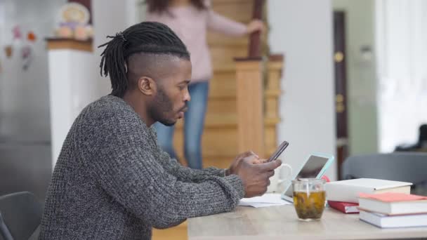 Sisi tampilan potret terkonsentrasi Afrika Amerika pria surfing online pesan internet sebagai gadis remaja gembira berjalan di bawah memeluk ayah berbicara. Kerja jauh dan gaya hidup keluarga. — Stok Video