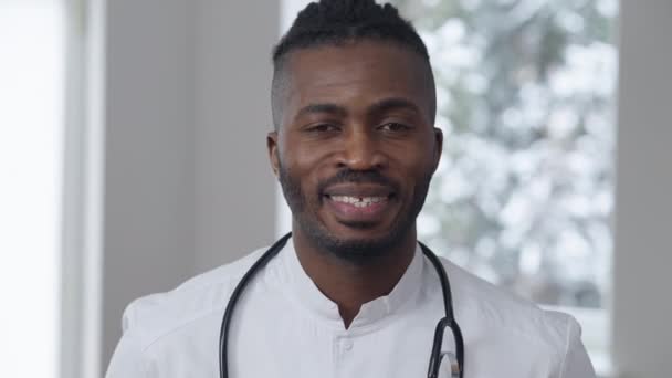 Close-up retrato de sorrindo positivo Africano americano médico do sexo masculino posando dentro de casa. Homem bonito alegre em uniforme médico olhando para a câmera em pé no hospital. Medicina e ocupação. — Vídeo de Stock