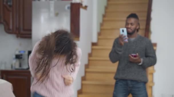 Média foto de despreocupado alegre menina adolescente afro-americana se divertindo dançando na sala de estar com o homem borrado sorridente filmando no smartphone em pé no fundo. Família positiva dentro de casa. — Vídeo de Stock