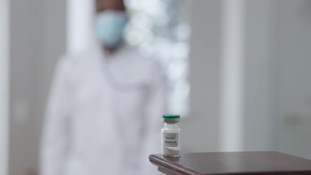 Covid-19 vacuna jab en la mesa con borrosa médico afroamericano caminando de fondo tomando vial y dejando. Médico irreconocible con tratamiento pandémico de coronavirus en interiores. — Vídeos de Stock