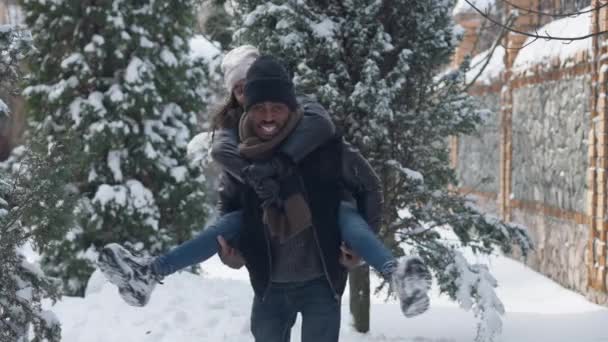 Mittlere Aufnahme eines fröhlichen afroamerikanischen Vaters, der seine Tochter auf dem Rücken hält und lächelnd in die Kamera blickt, die an einem verschneiten Wintertag im Freien steht. Glücklich mann und teen mädchen posiert auf hinterhof. — Stockvideo