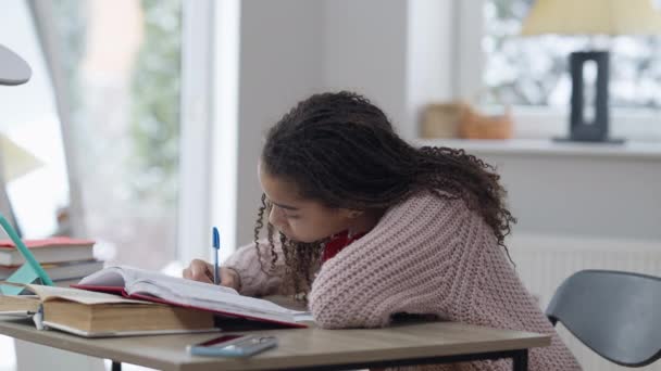 Diligente chica afroamericana que estudia en línea haciendo deberes en casa en interiores. Retrato de vista lateral de una colegiala inteligente inteligente inteligente escribiendo en un libro de trabajo sentado con una tableta y un libro. Educación. — Vídeo de stock