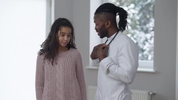 Portret van een positieve Afro-Amerikaanse dokter en tiener patiënt die naar de camera kijkt terwijl hij binnen staat te lachen. Vrolijke man en meisje poseren in het ziekenhuis. Geneesmiddelenconcept. — Stockvideo