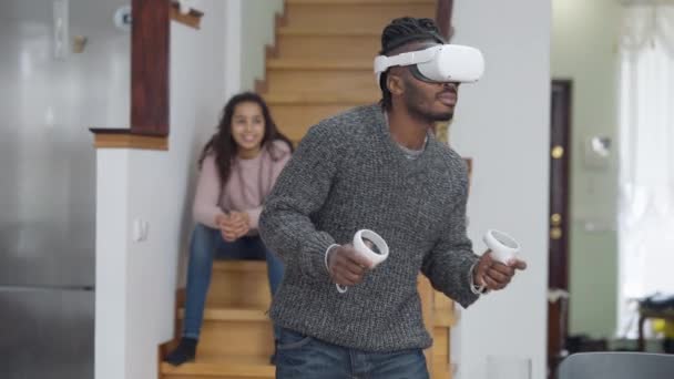 Hombre alegre en auriculares VR jugando en realidad aumentada como chica borrosa sentada en el fondo en las escaleras sonriendo. Mediana toma de positivo relajado afroamericano padre e hija en casa el fin de semana — Vídeo de stock