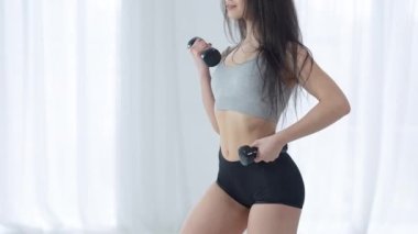 Tanınmayacak kadar ince, muhteşem Orta Doğulu kadın içeride duran dambılları kaldırıyor. Kendine güvenen genç sporcu kadın evde çalışıyor ve kameraya gülümsüyor. Spor ve sağlıklı yaşam tarzı.