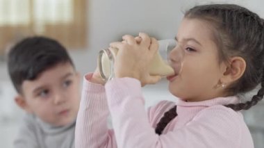 Neşeli Ortadoğulu kız muzlu kokteyl içiyor ve gülümsüyor. Bulanık erkek kardeş arka plandaki kız kardeşe bakıyor. Çocuklar evde mutfakta sağlıklı yiyeceklerin tadını çıkarıyorlar..