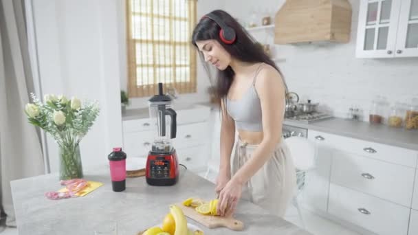 Giovane donna mediorientale in cuffia che balla in cucina affettando e mangiando limone. Ritratto di millenario positivo che si gode la frutta vitaminica a casa sorridendo. Hobby e stile di vita. — Video Stock