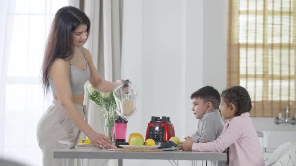 Mulher do Oriente Médio concentrada derramando smoothie em vidro preparando bebida de frutas saudável para crianças na cozinha. Filho e filha sentados na bancada à espera de um cocktail. Conceito matinal. — Vídeo de Stock