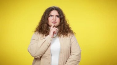 Büyük boy beyaz esmer, kıvırcık saçlı kadın kameraya gülümseyen bir fikir jesti yaptığımı gösteriyor. Sarı arka planda poz veren şişman, mutlu genç bayanın portresi. Vücut pozitif.