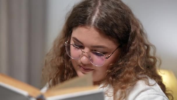 Detailní záběr mladé ženy v brýlích, jak čte knihu s úsměvem. Uvolněný šťastný bělošský čtenář si večer užívá koníčka doma. Koncept životního stylu a radosti. — Stock video