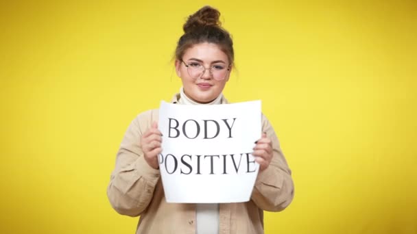 Selbstbewusste Frau mit Brille, die den Körper öffnet und in die Kamera lächelt. Porträt einer übergewichtigen jungen Kaukasierin mit Dutt-Frisur, die auf gelbem Hintergrund mit Banner posiert. — Stockvideo