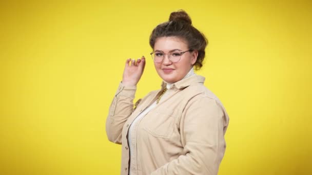 Mujer segura de tamaño grande positiva con peinado de moño en gafas girando a la cámara sonriendo. Retrato de alegre dama blanca con estilo posando sobre fondo amarillo. Confianza y estilo de vida. — Vídeo de stock