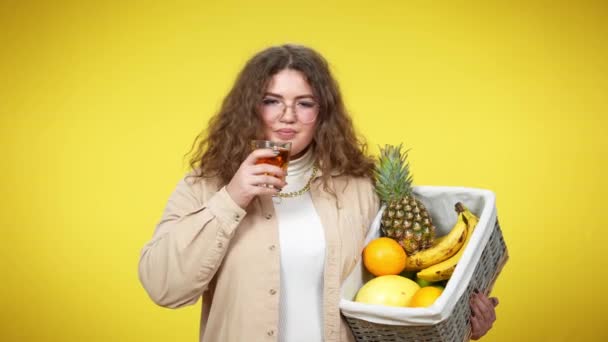 Jeune femme brune en surpoids dans les lunettes de boire du jus de grillage avec caméra tenant des fruits biologiques dans le panier. Portrait de sourire heureuse dame caucasienne posant à fond jaune. — Video