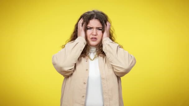 Mujer cansada de talla grande sosteniendo la cabeza en las manos mirando a la cámara con expresión facial triste. Retrato de una cansada mujer caucásica exhausta con migraña posando sobre fondo amarillo. Asistencia sanitaria. — Vídeos de Stock