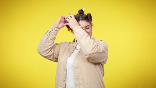 Mujer joven con sobrepeso aplicando rulos para el cabello mirando a la cámara. Retrato de dama milenaria caucásica de tamaño grande usando rodillos para el cabello haciendo peinado en fondo amarillo. Feminidad y belleza. — Vídeos de Stock