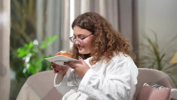 Sourire femme en surpoids sentant et mangeant savoureux croissant sucré. Portrait de confiant millénium caucasien plus-taille dégustant délicieux dessert dans la soirée à la maison. Mauvaise alimentation et obésité — Video