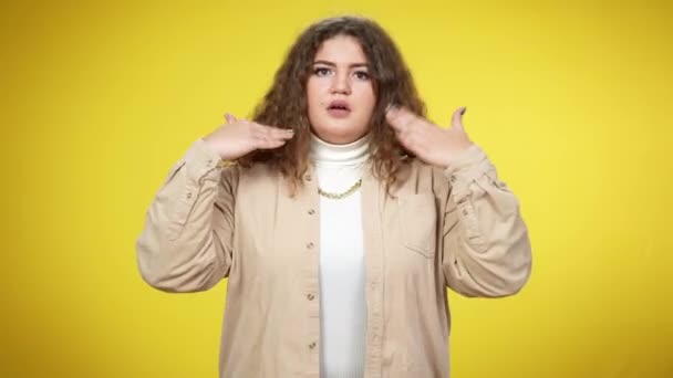 Portret van een jonge vrouw met overgewicht die naar een camera kijkt die handen schudt. Medium shot van plus-size geschokte blanke dame poseren op gele achtergrond. Panikeren en kalmeren. — Stockvideo