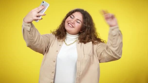 Alegre mujer millennial de tamaño grande tomando selfie en el teléfono inteligente en el fondo amarillo. Dispositivo adicto joven encantador sobrepeso dama caucásica fotografiando y sonriendo. Estilo de vida y adicción. — Vídeos de Stock