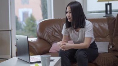 Neşeli Asyalı bin yıllık kadın dizüstü bilgisayarında görüntülü konuşma yapıyor rahat koltukta oturup konuşuyor ve gülümsüyor. Evde dinlenen ve internette sohbet eden pozitif ve rahat bir bayan. Modern yaşam tarzı.