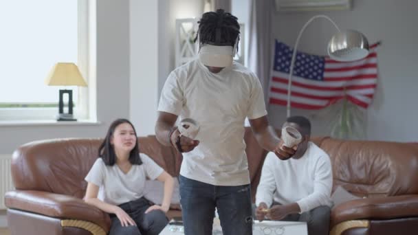 Lachende zelfverzekerde man in VR-headset boksen schaduw in augmented reality met vrienden juichen op de achtergrond. Vreugdevolle Aziatische en Afro-Amerikaanse jongeren die rusten en plezier hebben. Moderne technologieën. — Stockvideo