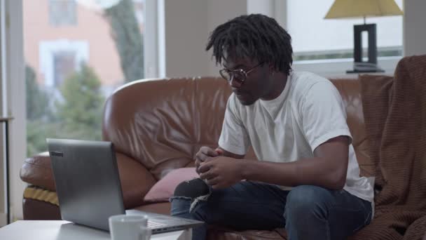 Jovem e bonito homem confiante conversando usando vídeo chat no laptop sentado em sofá confortável na sala de estar. Retrato de positivo afro-americano sorrindo millennial bate-papo on-line de casa. — Vídeo de Stock