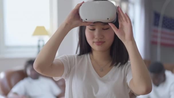 Positieve Aziatische vrouw die VR-headset afdoet met duim omhoog kijkend naar de camera als wazige Afro-Amerikaanse mannen die op de achtergrond op de bank zitten. Poftrait van opgewonden vrouwelijke gamer poseren binnen. — Stockvideo