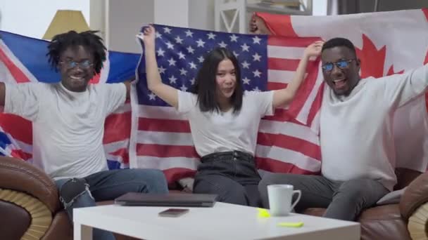 Amigos alegres posando con banderas dentro sentados en un cómodo sofá en la sala de estar. Mujer asiática sonriente y hombres afroamericanos hablando sacudiendo símbolos nacionales. Amistad internacional. — Vídeo de stock