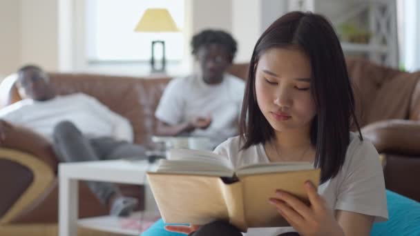 Retrato de vista frontal de la mujer asiática hermosa inteligente absorbida leyendo libro con hombres afroamericanos borrosos descansando en el fondo. Encantadora jovencita disfrutando de hobby en fin de semana en interiores. — Vídeos de Stock