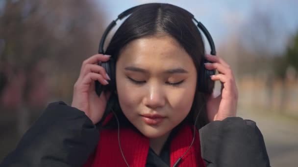 Nært ansikt til munter asiatisk ung kvinne som tar av seg hodetelefoner og smiler til kameraet i sakte film. Hodeskudd av gledelig, positiv tusenårsstilling utendørs på solskinnsdag. – stockvideo