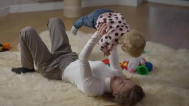 Çekici, zayıf, muhteşem bir milenyum annesinin kucağında çocuğu tutan bebekle konuşmasının yüksek açılı görüntüsü. Geniş açılı mutlu beyaz kadın portresi. Kızı yerde yatarken eğleniyor..