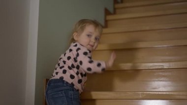 Yavaş çekimde el sallayan, merdivenlerden tırmanırken kameraya bakan sevimli, beyaz, sarışın kız. Evde poz veren güzel bebek portresi. Çocukluk ve mutluluk.