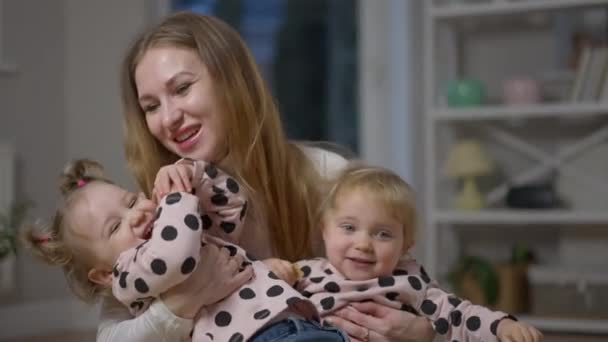 Ritratto di madre caucasica felice abbracciando e baciando figlie gemelle. Sorridente bella donna con bambine a casa al chiuso. Maternità e felicità concetto. — Video Stock