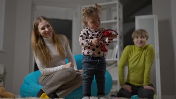 Široký záběr portrét hezké holčičky, jak si nasazuje sluchátka stojící v obývacím pokoji s matkou a babičkou obdivující dítě z pozadí. Roztomilé dítě baví hraní doma. — Stock video
