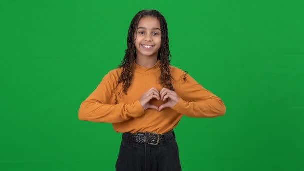 Mutlu güzel Afro-Amerikalı genç kız kameraya gülümserken kalp hareketi gösteriyor. Yeşil ekran kromatonunda poz veren kendinden emin ergen bir gencin portresi. Aşk konsepti. — Stok video