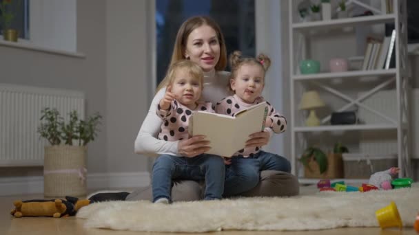 Brett fotograferat porträtt av unga kaukasiska mor läser bok för tvillingdöttrar sitter i vardagsrummet. Glad charmig kvinna och små flickor njuter av familjen fritid hemma. Långsamma rörelser. — Stockvideo