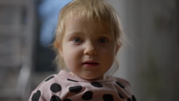 Close-up da linda criança bebê caucasiano com cabelo loiro olhando ao redor e para a câmera. Retrato da menina encantadora bonito dentro de casa. Beleza e infância. — Vídeo de Stock