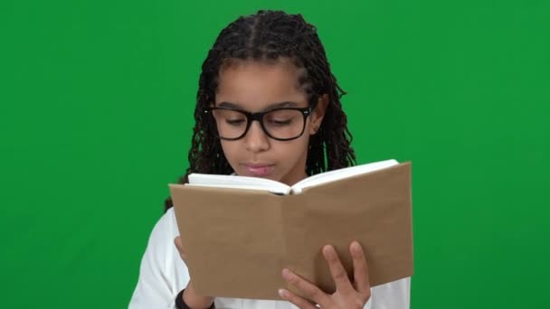 Encantadora adolescente afroamericana con gafas leyendo libro en pantalla verde mirando a la cámara sonriendo. Retrato de inteligente inteligente hermosa adolescente posando cromakey. Educación e inteligencia. — Vídeo de stock