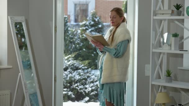 Inteligentna stylowa kobieta w średnim wieku czyta książkę stojącą przy szklanych drzwiach w luksusowym domu. Widok z boku portret eleganckiej białej pani korzystających hobby w domu w weekend. Bogactwo i styl życia. — Wideo stockowe