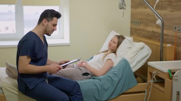 Side view portrait of smiling positive Middle Eastern male doctor sitting on hospital bed talking to female Caucasian patient. Young man supporting ill woman lying in clinic indoors. Slow motion. — Stock Video