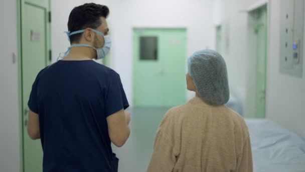 Vue arrière du médecin et du patient marchant vers la salle d'opération à la clinique parlant au ralenti. Caméra en direct suit professionnel Moyen-Orient homme et femme caucasienne se promener pour l'opération. — Video