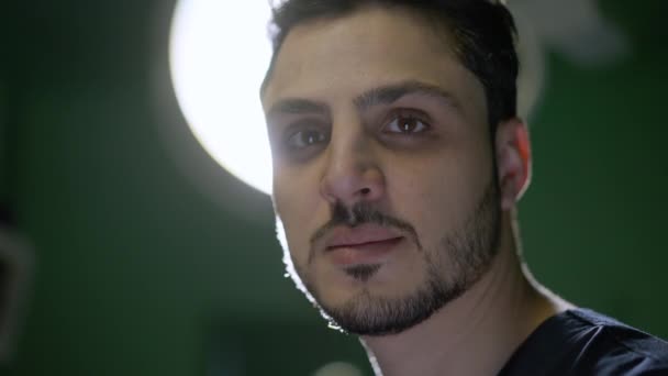 背景に輝く外科用ランプでカメラを見て若いハンサムな中東の男のクローズアップ顔。手術室でポーズをとっている重大な自信のあるプロの外科医のヘッドショットの肖像. — ストック動画