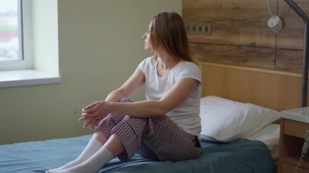 Genç üzgün bir kadının hastane yatağında oturup düşünmesi. Depresyondaki zayıf, güzel, beyaz hasta koğuşta. Hastalık ve tıp konsepti. Yavaş çekim. — Stok video