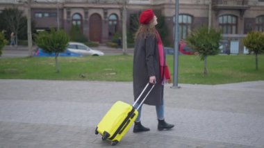 Side view wide shot of young confident elegant woman strolling along city street with yellow travel bag. Stylish Caucasian female tourist walking in urban town with luggage in slow motion.