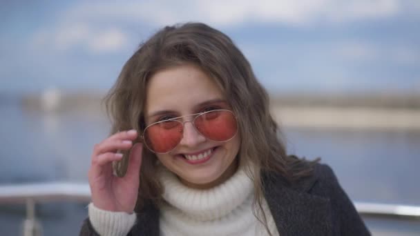 Közelkép egy vidám, gyönyörű, napszemüveges fiatal nőről, aki a szabadban álló kamerát nézi, ahogy mosolyog a napsütéses tavaszi őszi napon. Vidám, boldog, nyugodt évezred a városi töltésen. — Stock videók