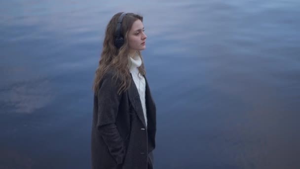 Seitenansicht einer eleganten, charmanten jungen Frau mit Kopfhörern, die auf einem Damm mit blauem Flusswasser im Hintergrund spaziert. Selbstbewusste schlanke stylische kaukasische Millennials gehen in Zeitlupe im Freien. — Stockvideo