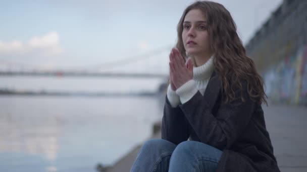 Hopeless lonely young woman holding hands together thinking sitting on urban city embankment looking away. Portrait of depressed Caucasian beautiful brunette suicidal millennial outdoors. — Stockvideo