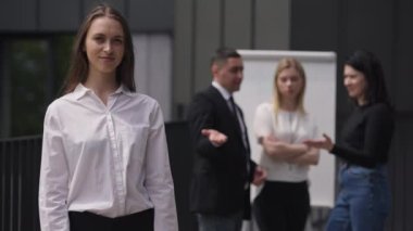 Kendine güvenen kibirli kadının portresi kameraya bakarken arka planda alay eden meslektaşlarını görmezden geliyor. Beyaz tenli genç, güzel bir çalışan ağır çekimde dışarıda poz veriyor..