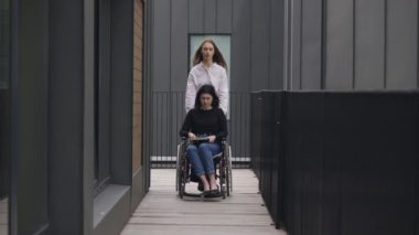 Wide shot portrait of young Caucasian woman pushing wheelchair with disabled colleague outdoors. Beautiful slim coworker taking care of paraplegic invalid. Support and inclusivity.