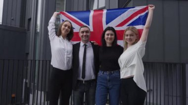 Four confident British business people raising national flag in slow motion looking at camera smiling. Group of friendly managers posing outdoors with national flag. Patriotism and business.