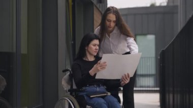 Disabled Caucasian CEO in wheelchair discussing business project with assistant outdoors. Serious successful women talking analyzing strategy. Slow motion.